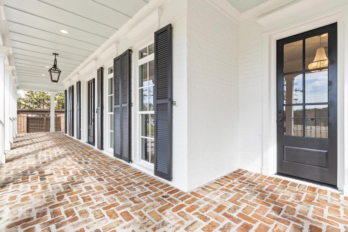 House plan exterior showing a covered porch with brick flooring, white columns, black shutters, and a black front door.