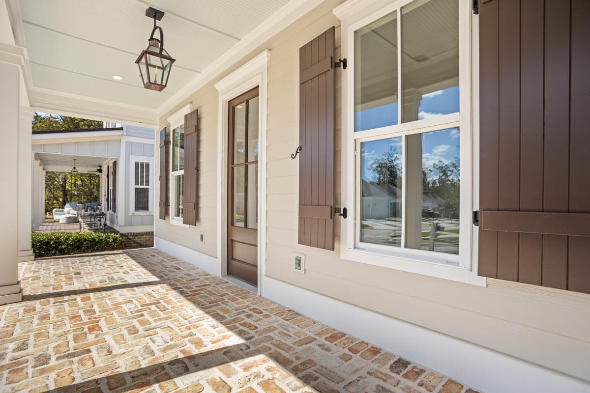 House plan exterior with a covered porch, brick pavers, paneled door, and louvered shutters. Traditional gas lantern hangs from ceiling.