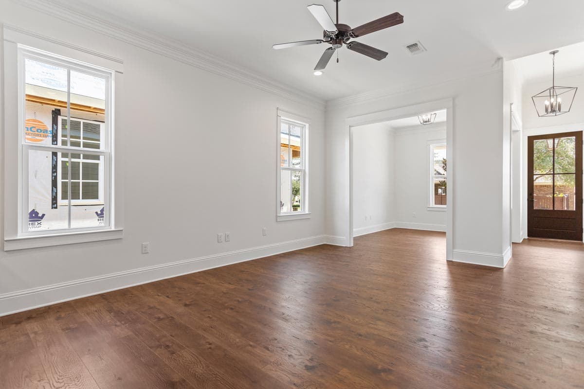 Interior view of an empty room with hardwood floors, white walls, a ceiling fan, and an open doorway to another room and the front entrance.