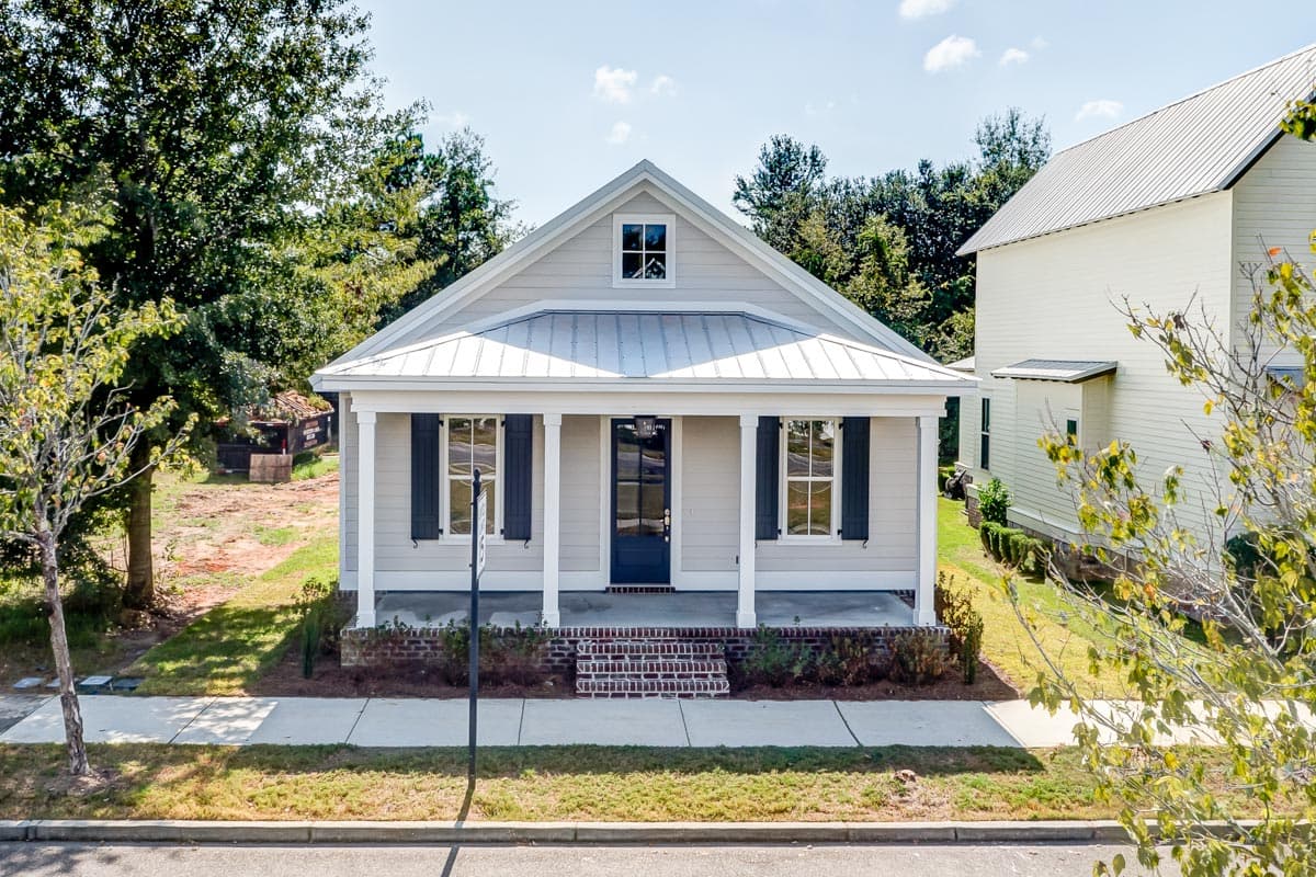 House plan exterior. One-story Cottage style with front porch, gabled roof, and dark shutters. Metal roof. Brick foundation.