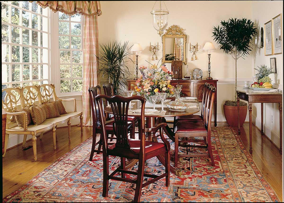 Dining room with a round table, chairs, ornate bench, large windows, sideboard, and patterned rug.