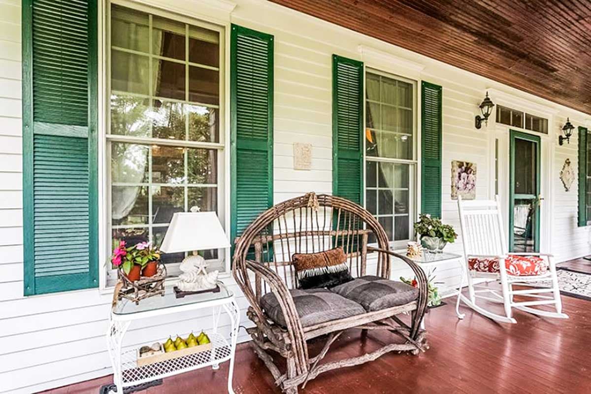 House plan exterior: two-story white house with green shutters, double-hung windows, and a covered porch with seating.