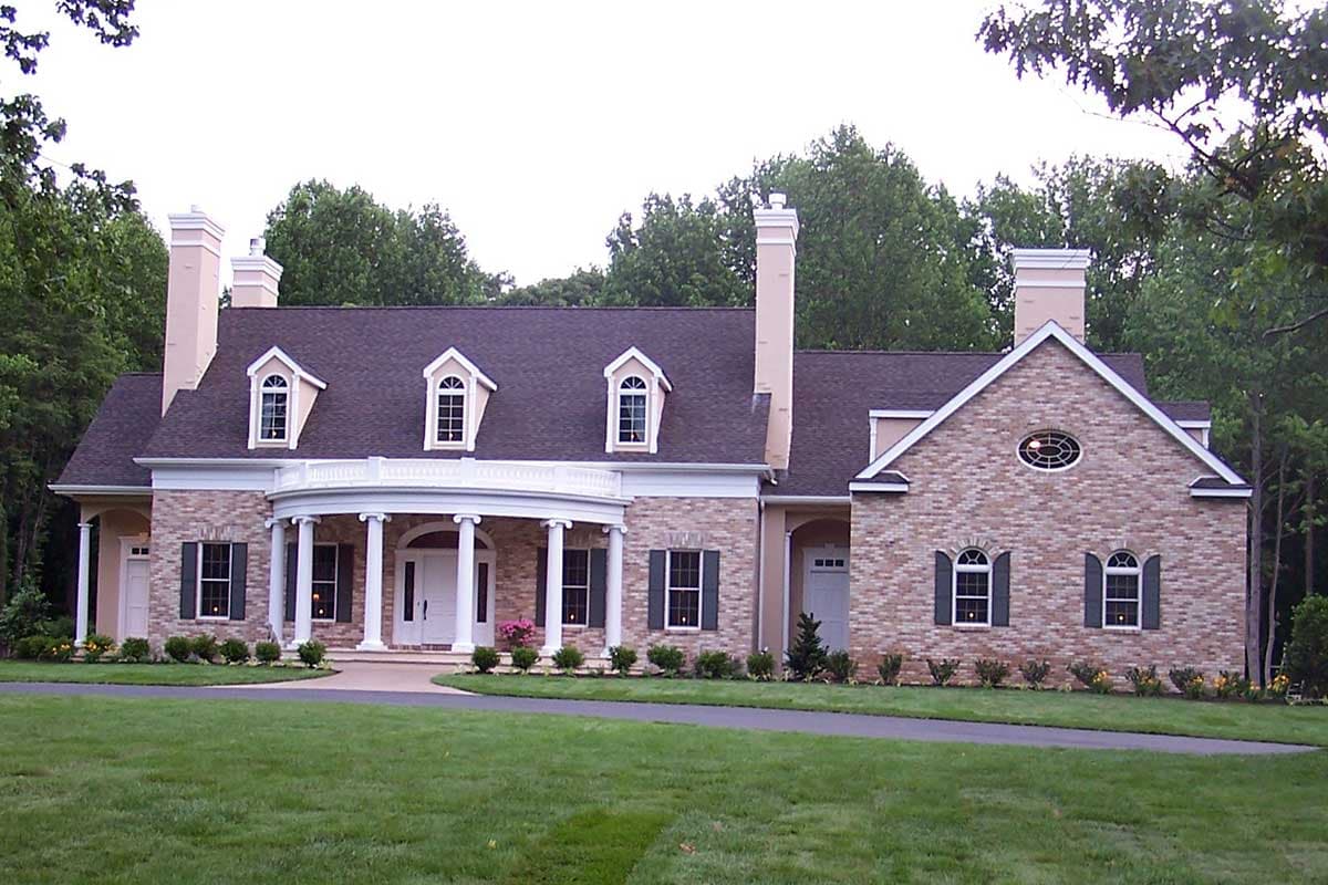 House plan exterior. Two-story Colonial with a wraparound porch supported by columns, dormers, and brick facade with arched window details.