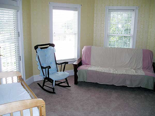 Interior view of a room with a rocking chair, futon, and crib. Two windows with blinds are visible.