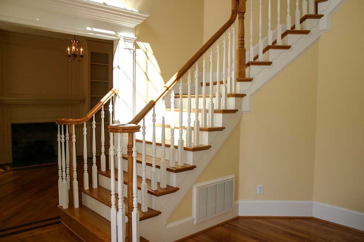 Interior view of staircase with wooden treads, white balusters, and carved newel posts. Sunlight streams through window.