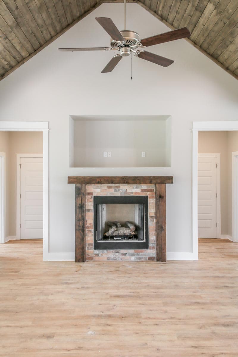 Interior view of a great room with a brick fireplace, vaulted wood-paneled ceiling, ceiling fan, and two doorways.