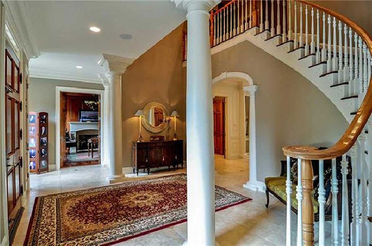 Interior view of a grand foyer with a curved staircase, tiled floor, ornate rug, and arched doorways.