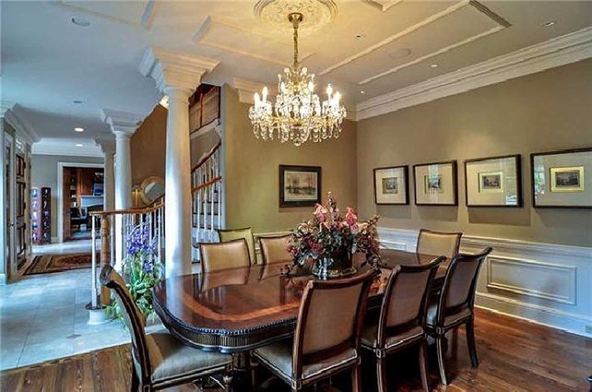 Dining room with a large wooden table, upholstered chairs, crystal chandelier, columns, and artwork.