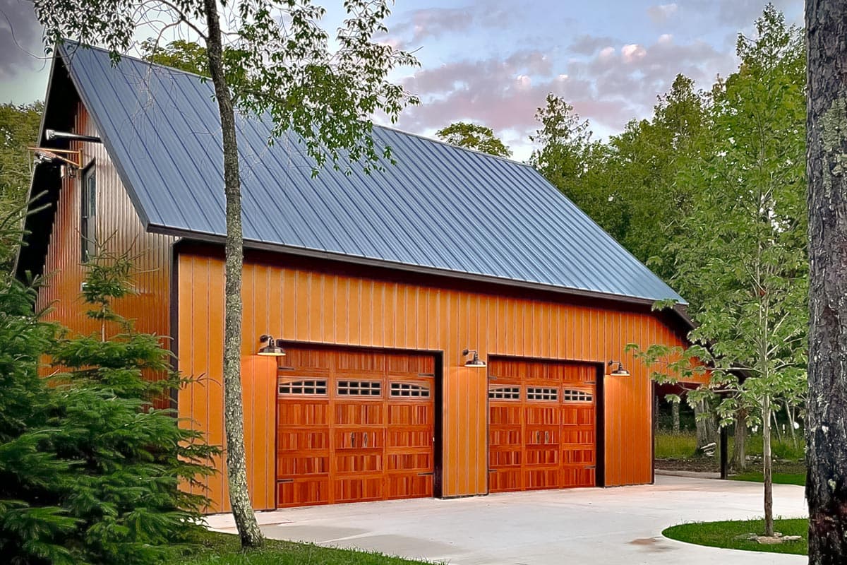 House plan exterior. Two-story structure with gable roof and metal siding. Features include two garage doors and exterior lighting.