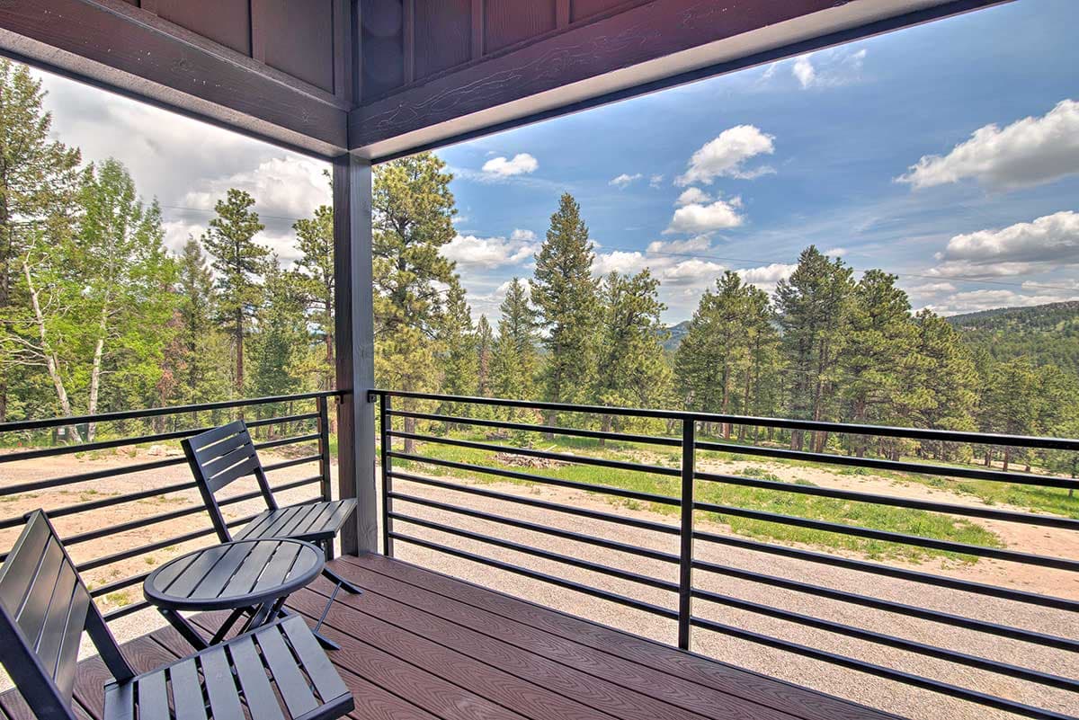 Covered porch with dark wood deck, metal railings, and seating overlooking a treed landscape under a cloudy sky.