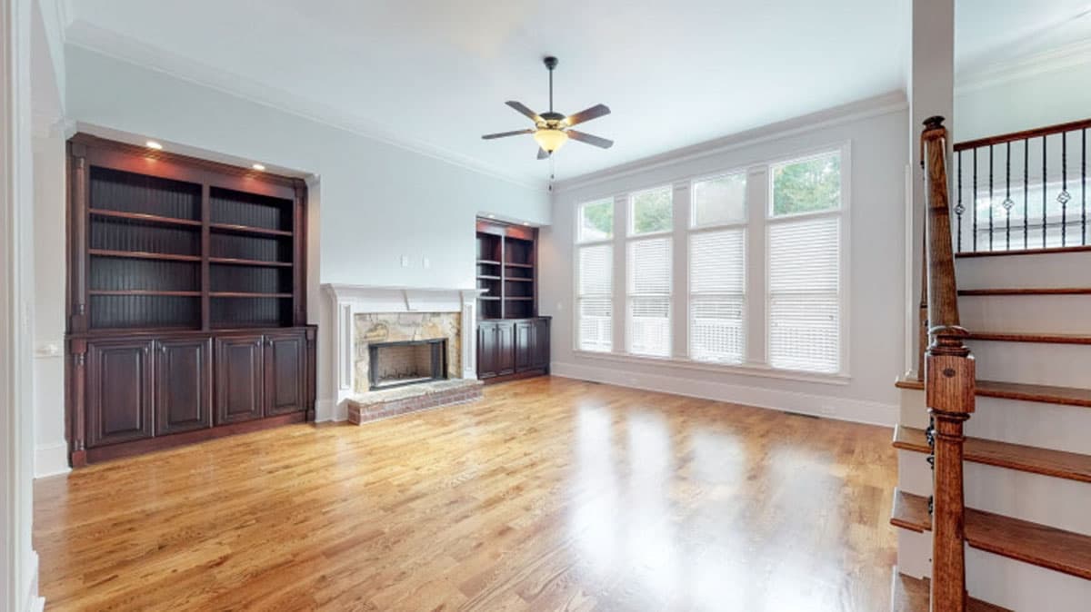 Living room with fireplace, built-in dark wood bookshelves, large windows, ceiling fan, and wood staircase.