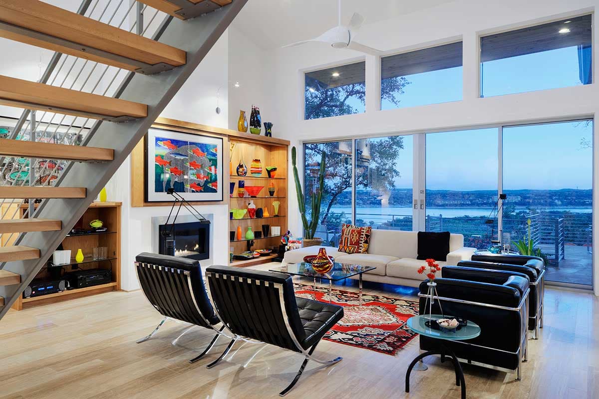 Living room with modern staircase, fireplace, floor-to-ceiling windows with lake view, and contemporary furniture.