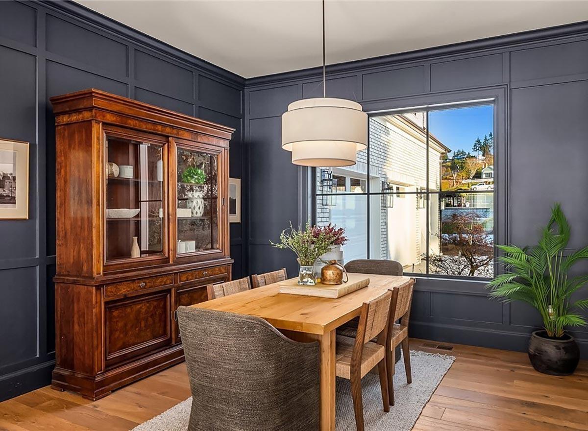 Dining room with wood table and chairs, paneled dark walls, glass-front china cabinet, and large grid window.