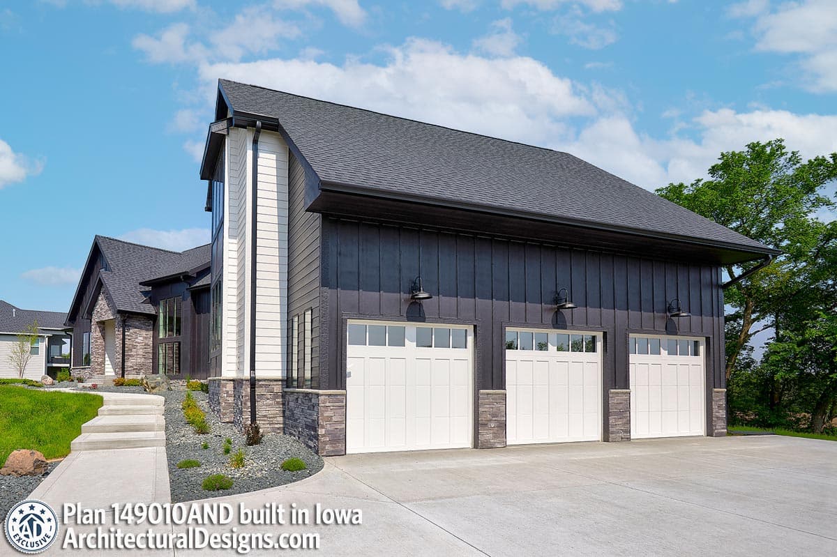 House plan exterior with three-car garage, dark vertical siding, stone accents, white garage doors, and gabled roof.