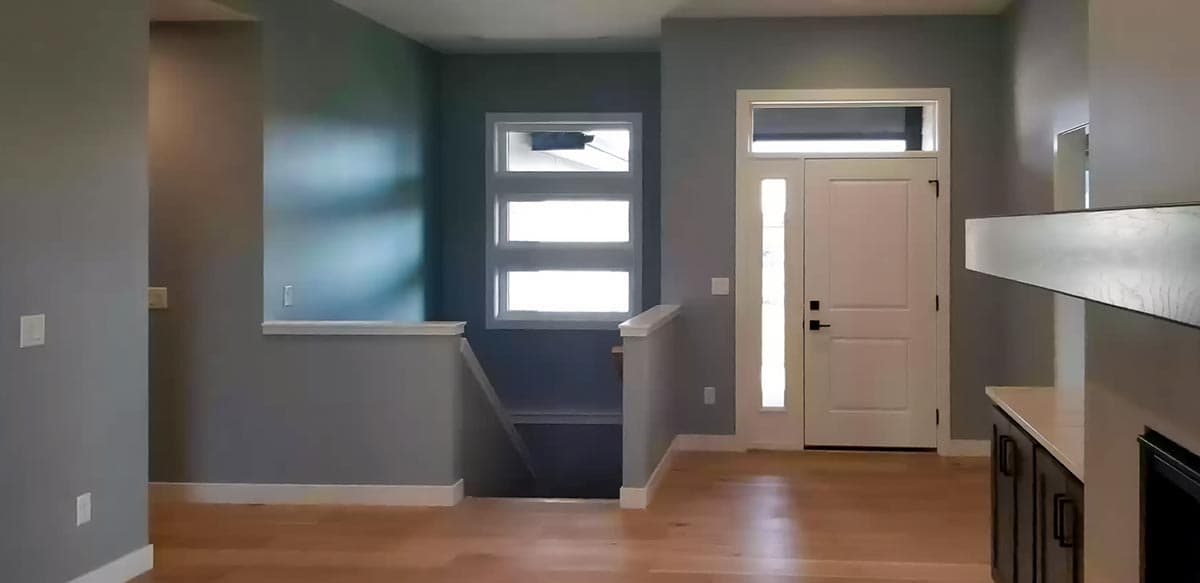 Interior view of a house showing a front door with sidelights, stacked windows above stairs, and a partial view of kitchen cabinetry.