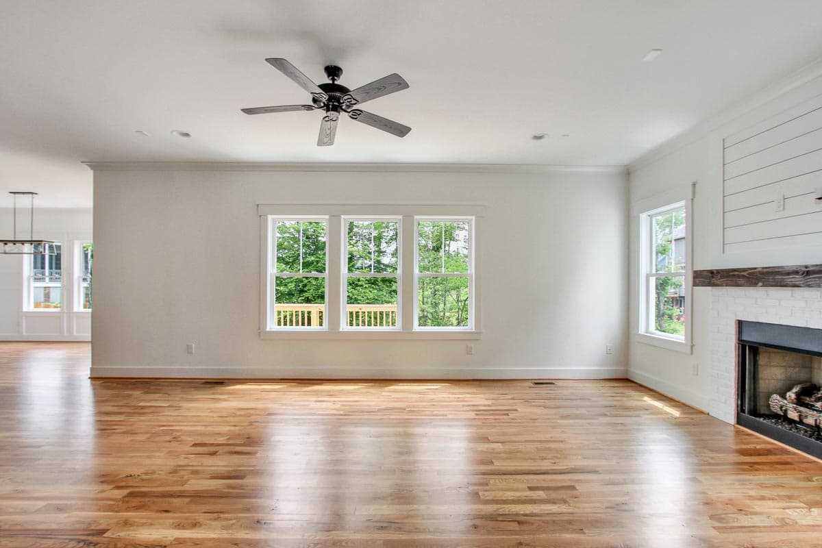 Interior view of a great room with hardwood floors, a ceiling fan, fireplace, and three windows overlooking trees.