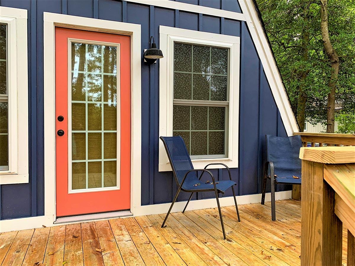 House plan exterior showing a navy blue vertical board and batten siding, a coral door with grid panes, and two windows on a wooden deck.