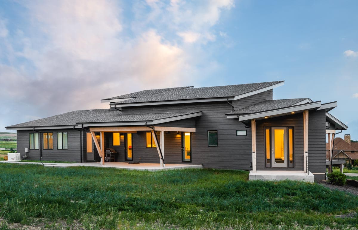 Modern house plan exterior with a low-pitched roofline, dark siding, covered patio with exposed beams, and prominent gables.