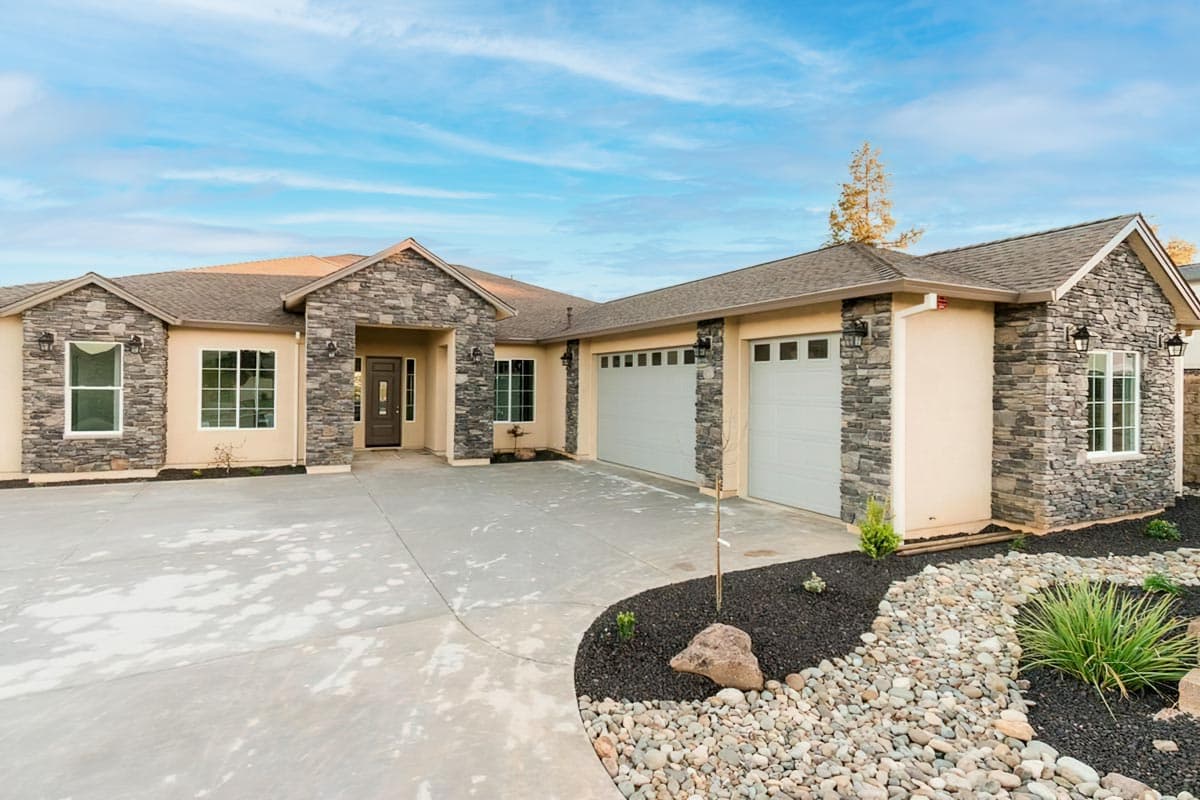 House plan exterior. One-story Ranch style home with stone accents, gables, and a three-car garage. Features a concrete driveway and landscaping.