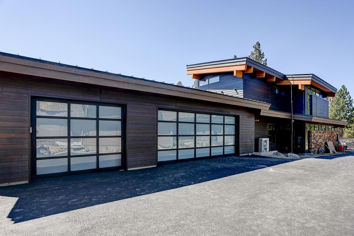 Modern house exterior with dark wood siding, two glass panel garage doors, and cantilevered roof sections.