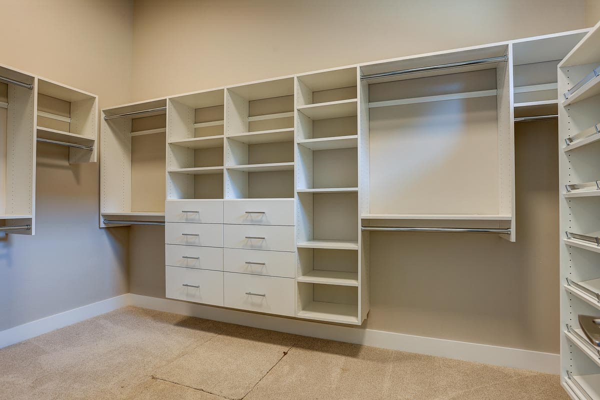 Walk-in closet with white shelving units, multiple drawers, and hanging rods against a neutral wall.