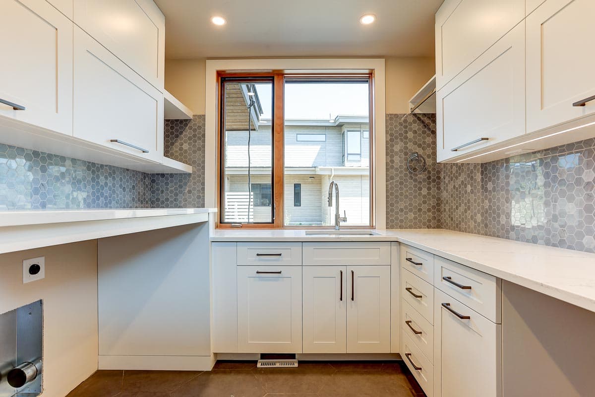 Laundry room with white cabinets, quartz countertops, gray hexagon tile backsplash, and a window overlooking houses.