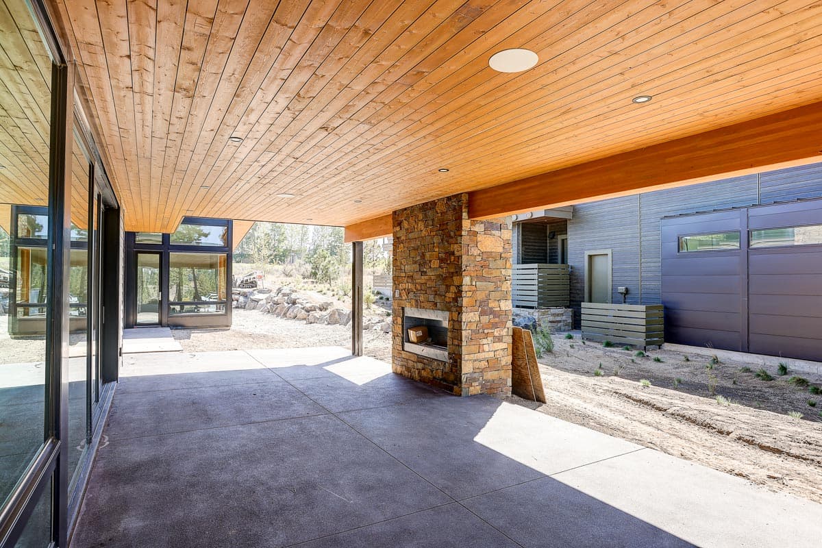 Covered patio with stone fireplace and wood plank ceiling. Modern home exterior with large windows and dark siding.