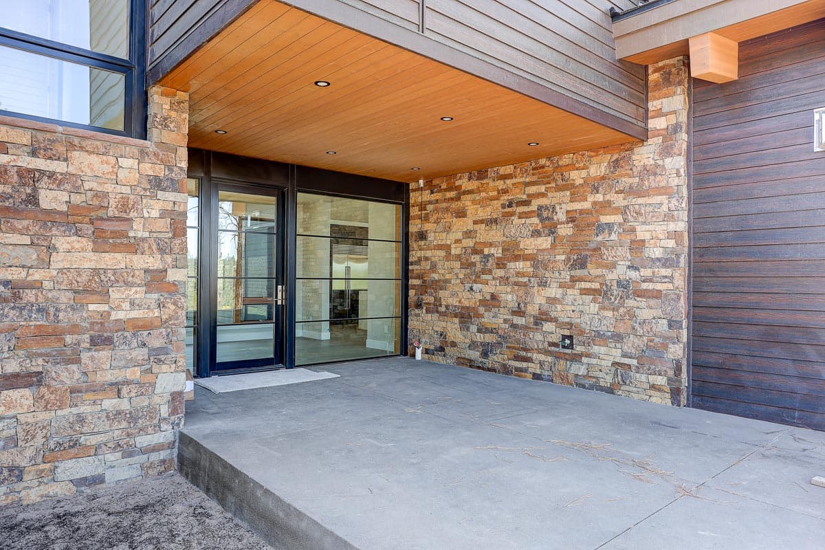 Modern house exterior with stacked stone, wood siding, and large glass entry doors under wood-paneled overhang.