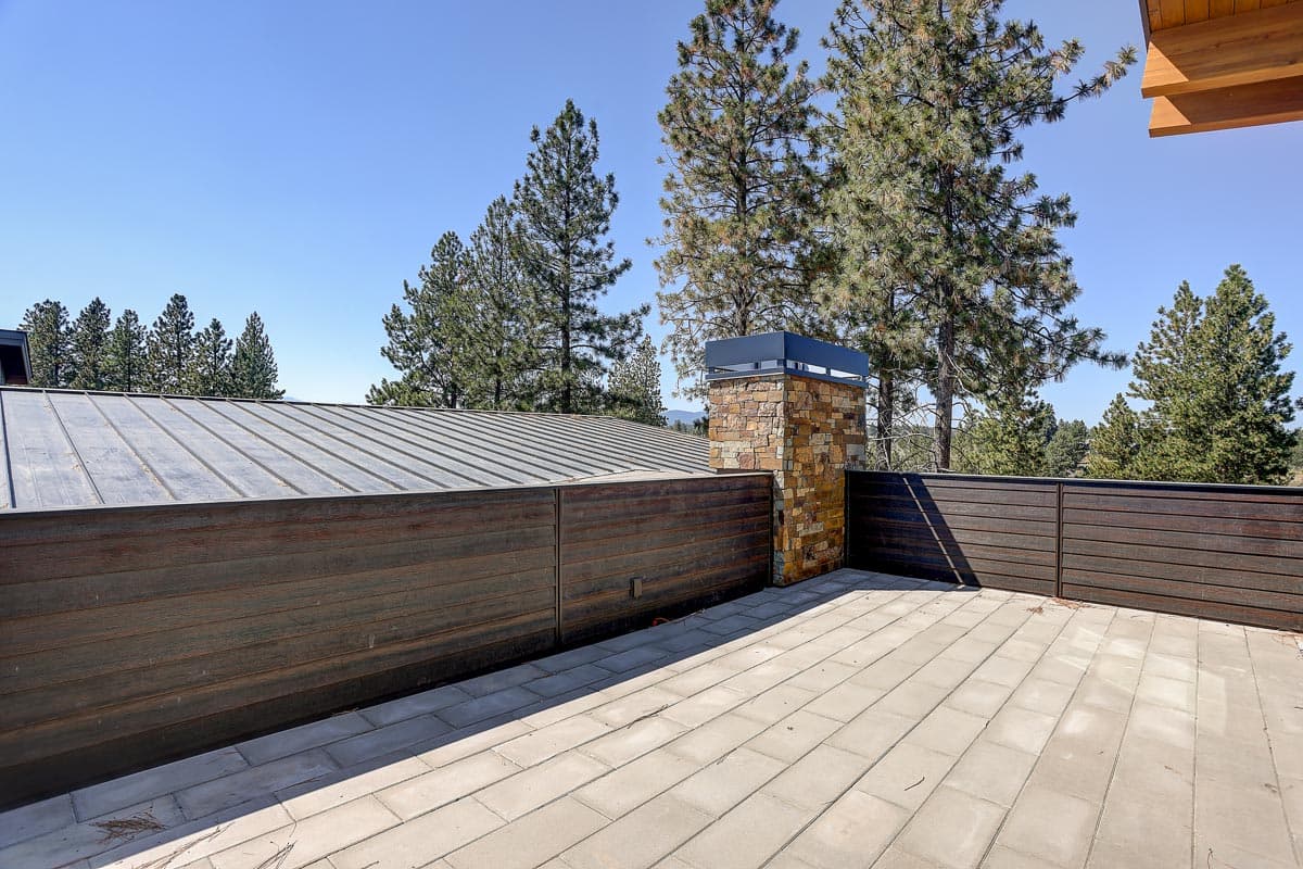 Modern house plan exterior with metal roof, stone chimney, horizontal wood siding fence, and paved patio.