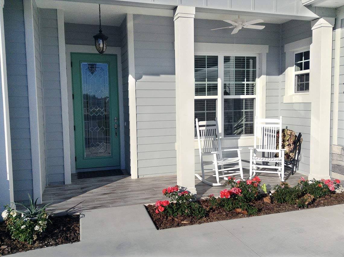 House plan exterior: Covered porch with two white rocking chairs, a teal front door with decorative glass, and double windows.