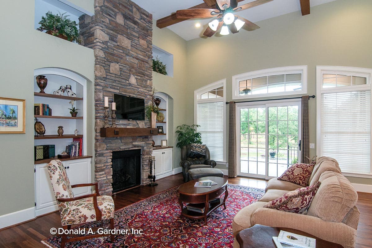 Great room with stone fireplace, built-in shelving, vaulted ceiling with fan, and large windows overlooking backyard.