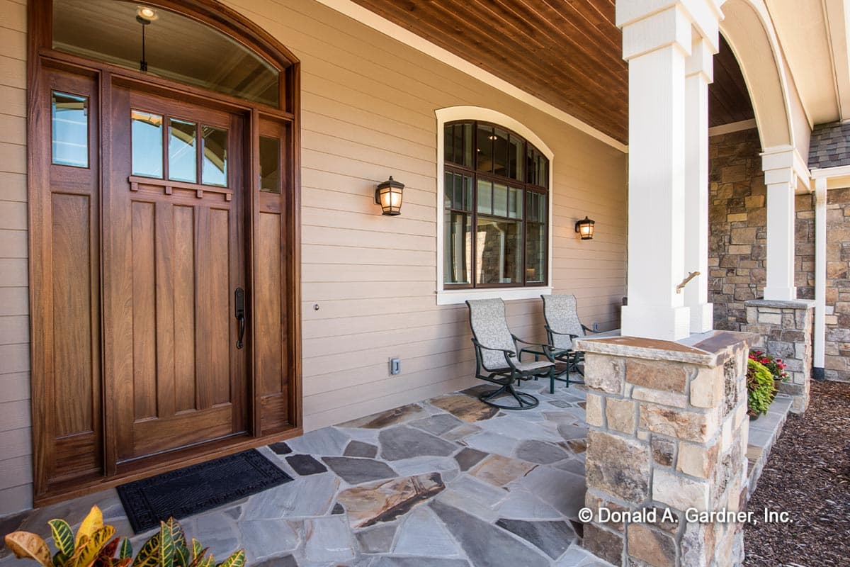 House plan exterior featuring a wooden arched entry door, stone-clad porch pillars, and large arched window.