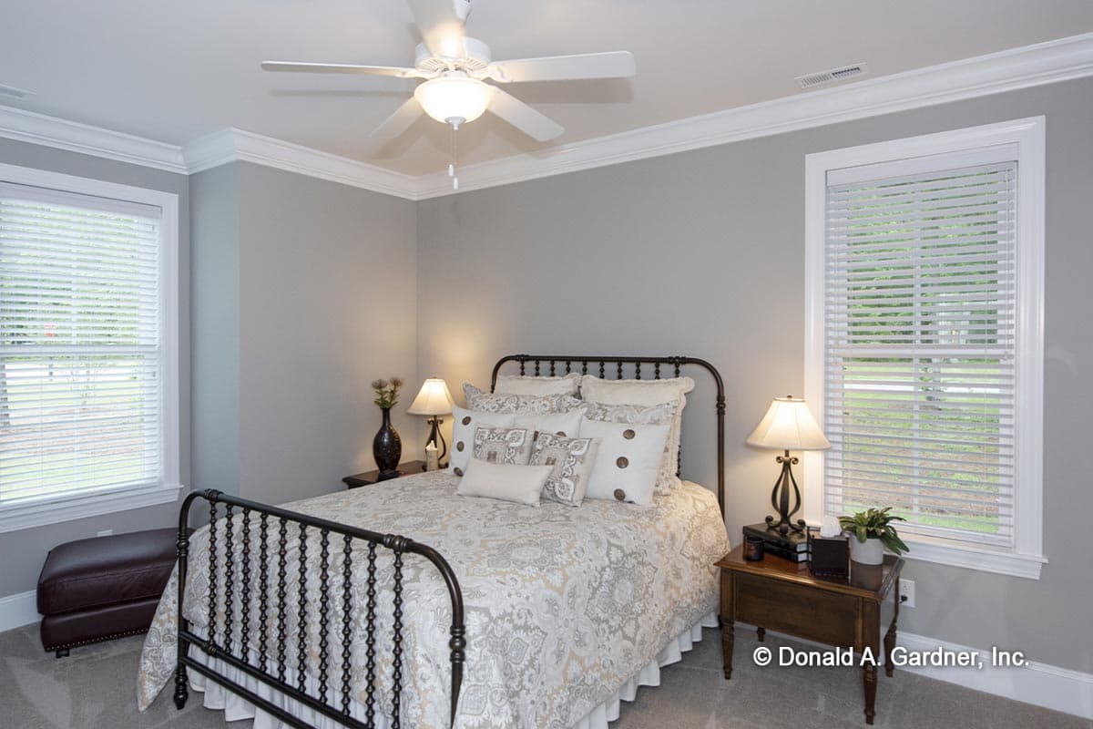 Bedroom interior with metal bed, decorative pillows, ceiling fan, two windows with blinds, and a leather ottoman.