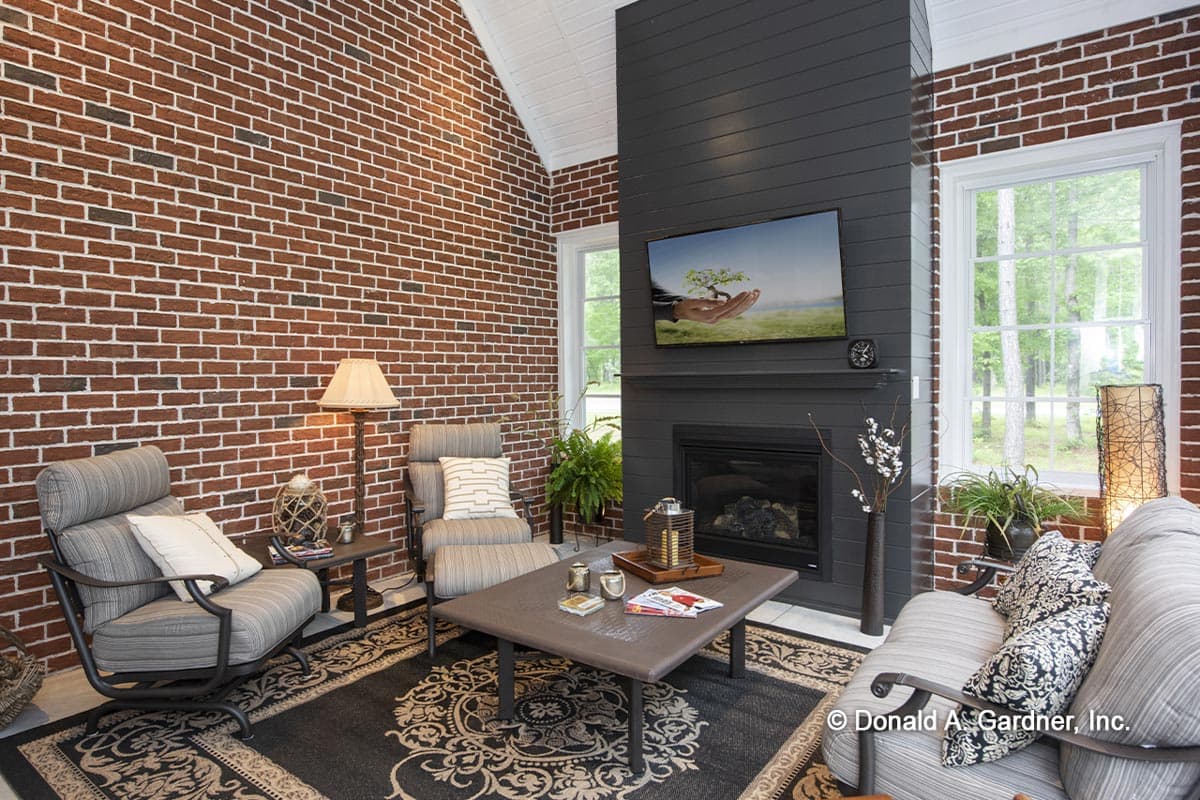 Living room with vaulted ceiling, brick accent wall, fireplace with mounted TV, and large windows.