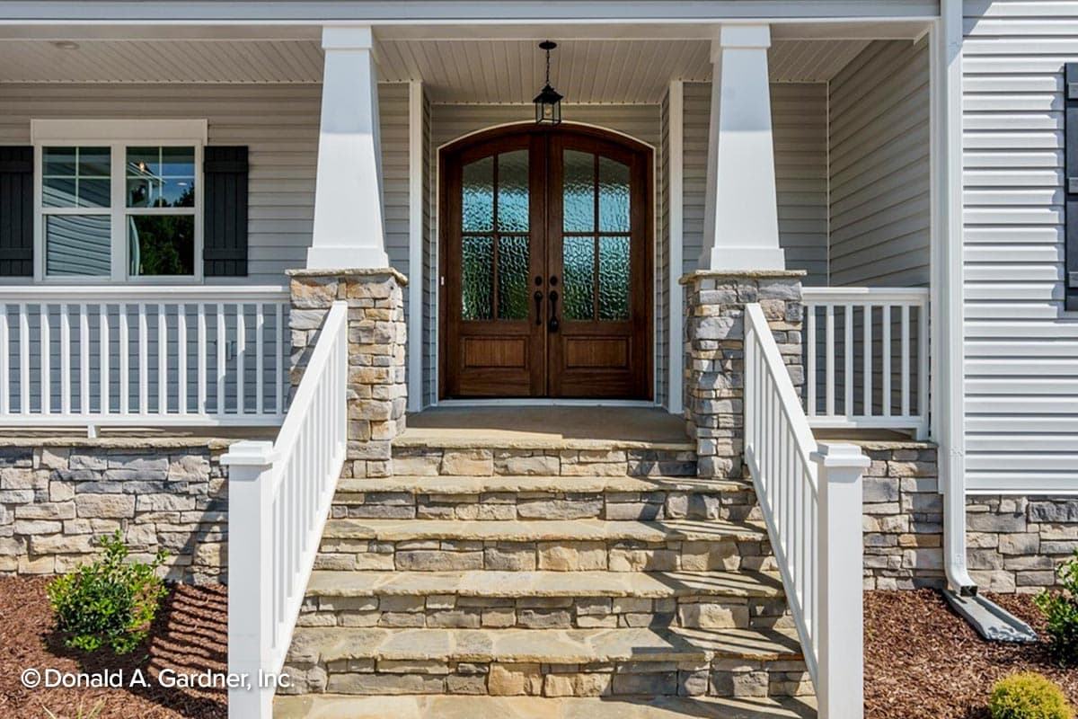 House plan exterior featuring stone-clad porch supports, double wooden doors with arched glass, and a white railing.