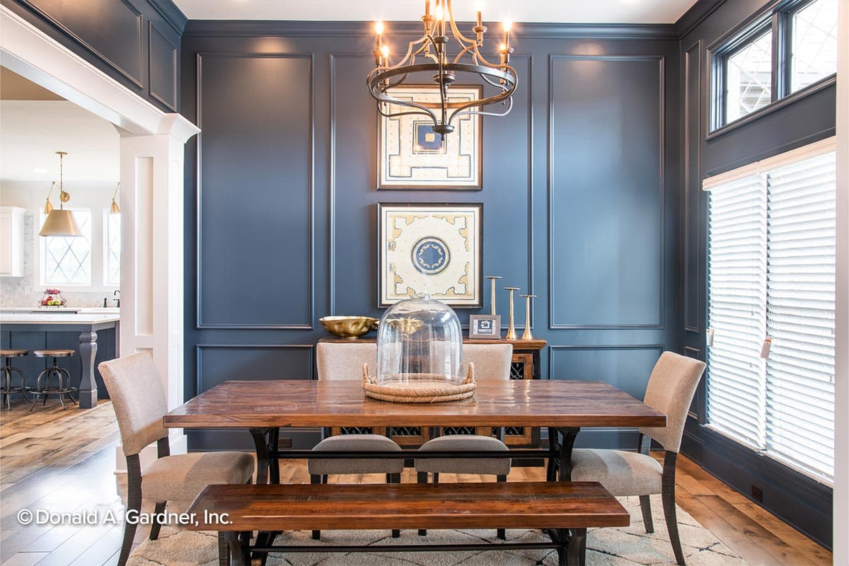 Dining room with paneled walls, a wood table and bench, and view into a kitchen with island and stools.