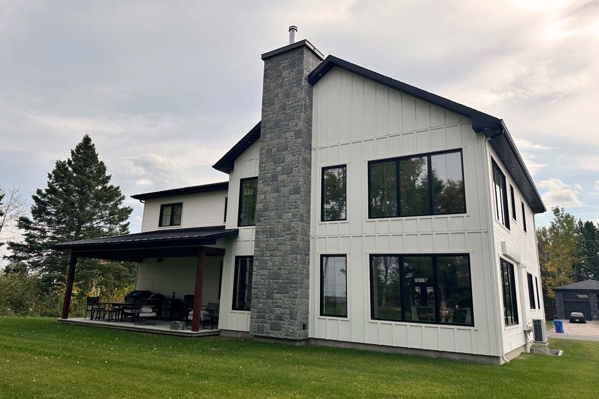 Modern two-story house with a prominent stone chimney, white siding, black-framed windows, and a covered patio overlooking a green lawn.