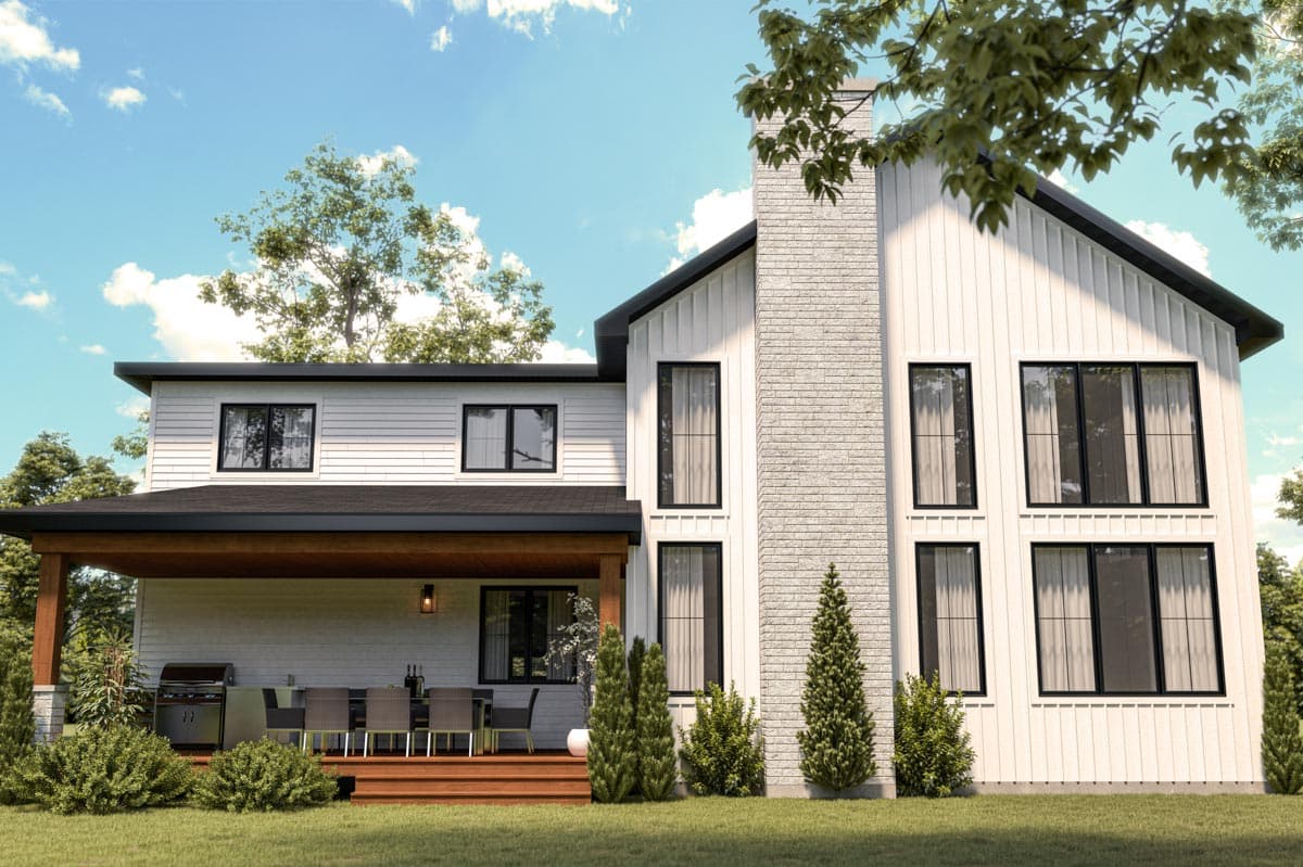 A modern two-story white house with black trim and a stone chimney. A wooden deck with outdoor dining faces the lawn with lush greenery.