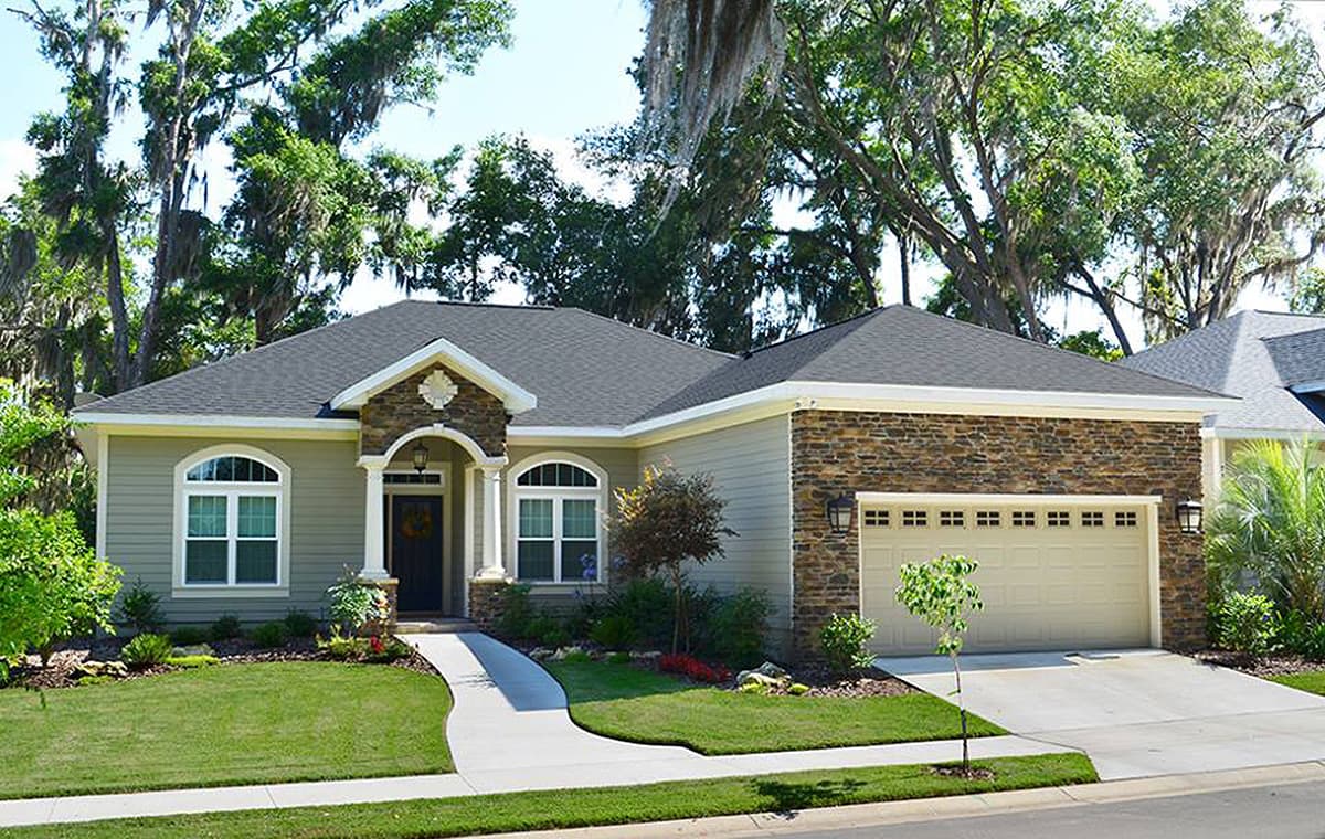 House plan exterior. Ranch-style home with stone facade on garage, arched entry, and gabled roof. Features include a walkway and mature landscaping.