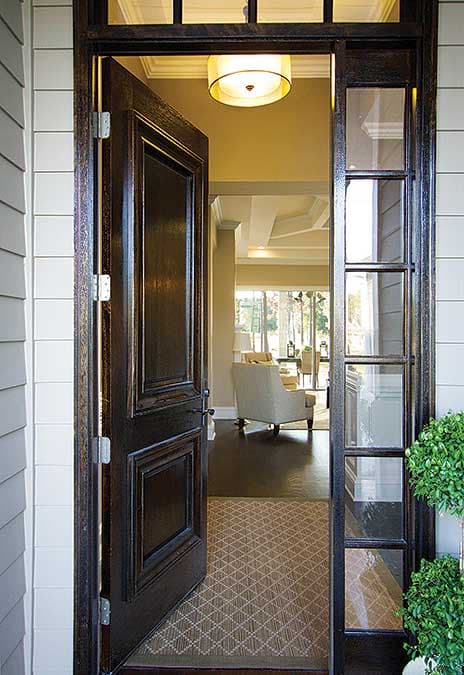 Dark wood paneled door open to foyer with tray ceiling, view to living room with furniture and large windows.