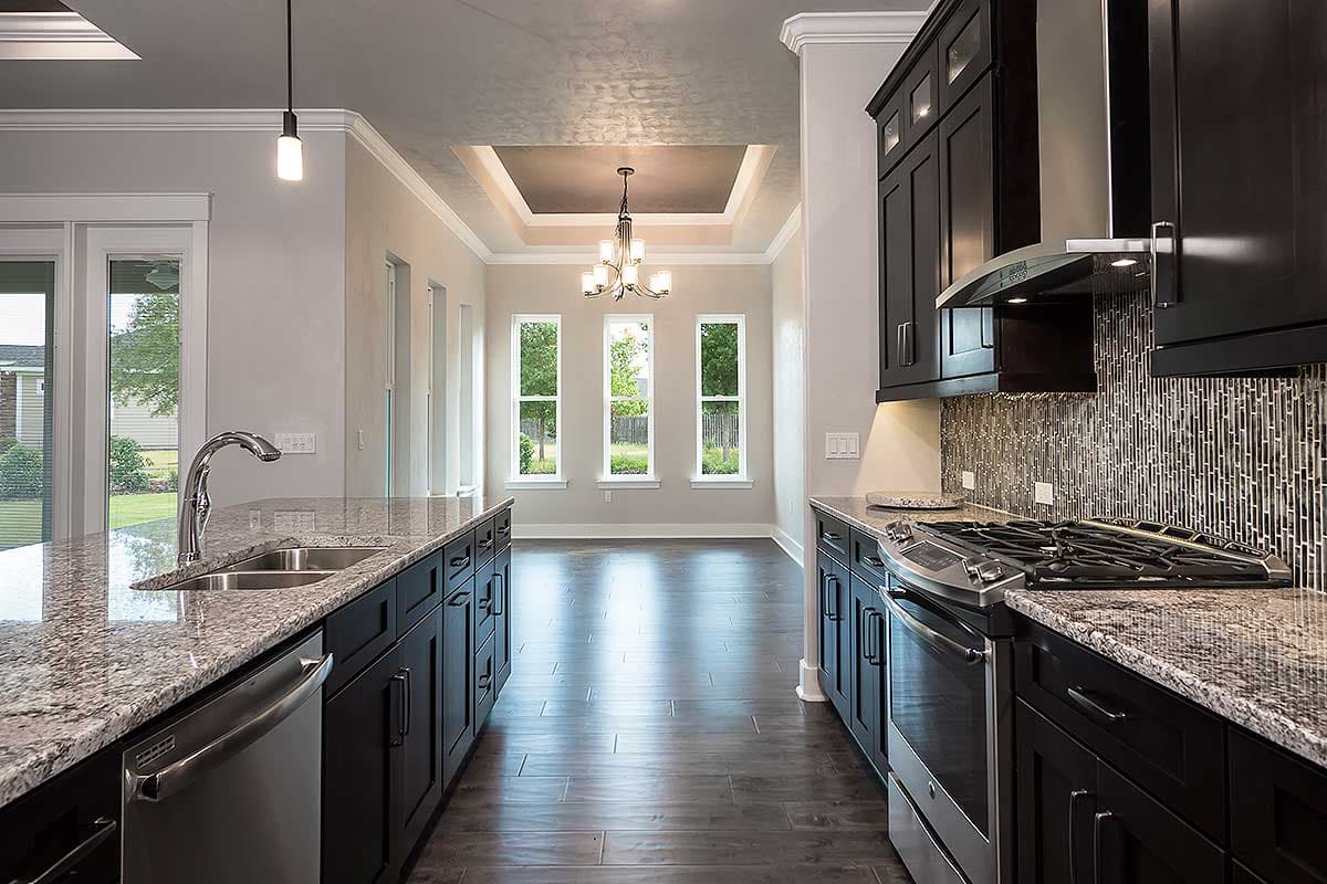 Kitchen with dark cabinetry, granite countertops, stainless steel appliances, and view into dining area with tray ceiling and three windows.