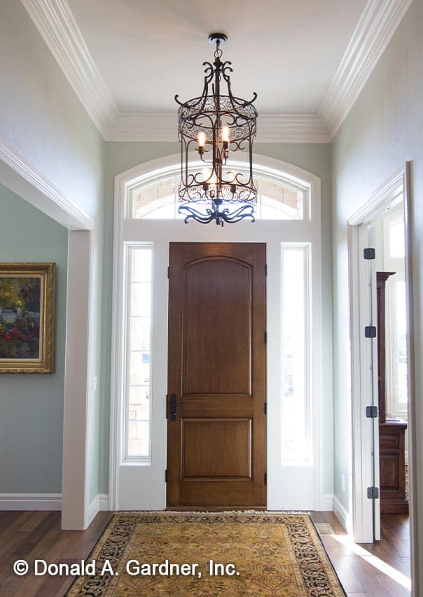 Interior view of a house entryway with a paneled wooden door, sidelights, arched transom window, and decorative chandelier.