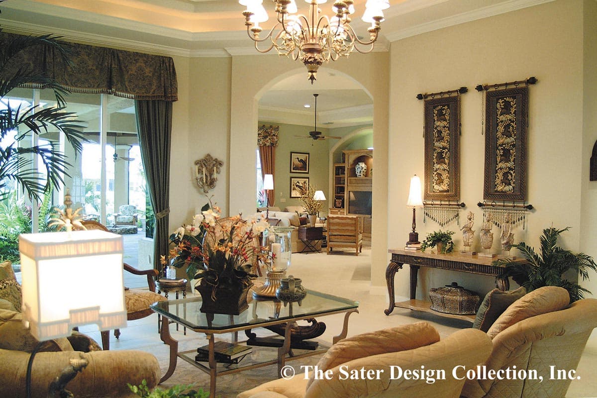 Interior view of a living room with an open archway to a family room, featuring a glass coffee table and ornate chandelier.