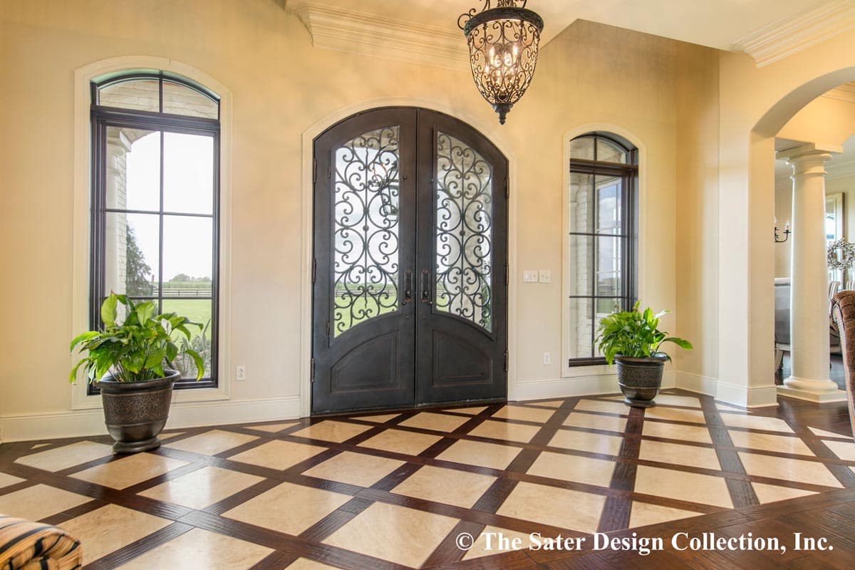 Interior view: Double arched entry doors with iron scrollwork, flanked by arched windows, leading to a foyer with patterned marble and wood flooring.