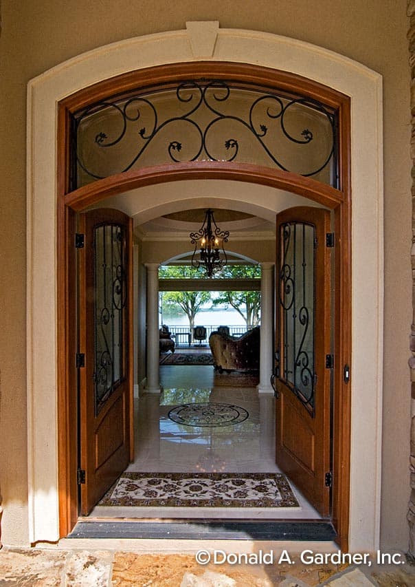 Exterior view of house plan featuring arched entry, double doors with wrought iron detail, and interior view of foyer with chandelier and lake view.