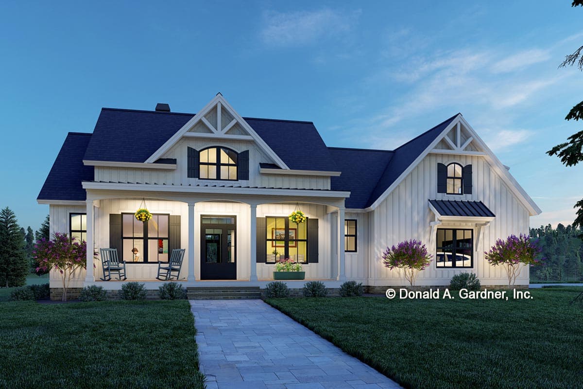 Exterior of a white farmhouse with dark roof and trim, featuring porch with rocking chairs and a walkway at dusk.
