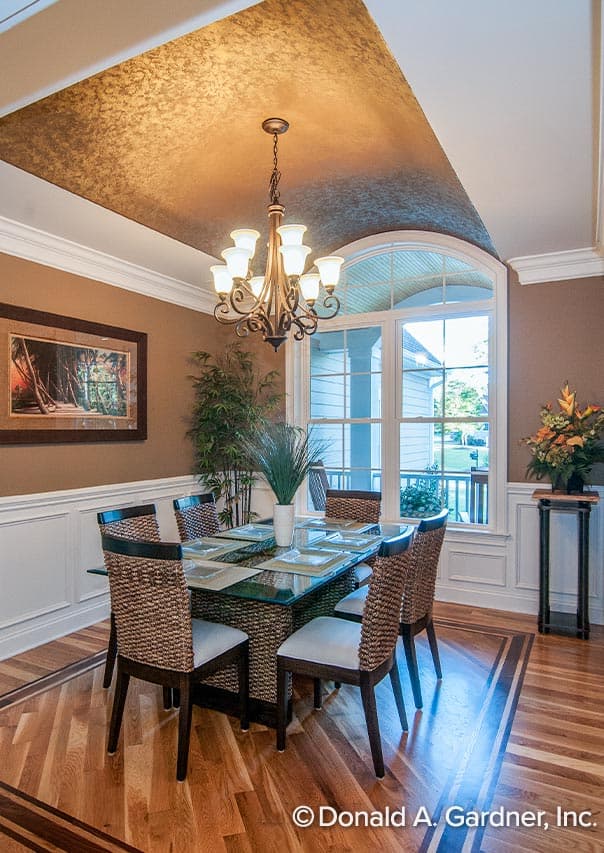 Dining room with tray ceiling, chandelier, glass-top table, wicker chairs, and arched window.