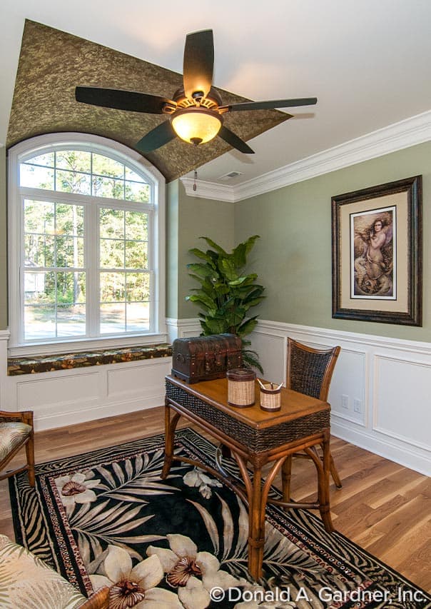 Interior view of a study with a large arched window, wainscoting, a rattan desk and chair, and a tropical rug.