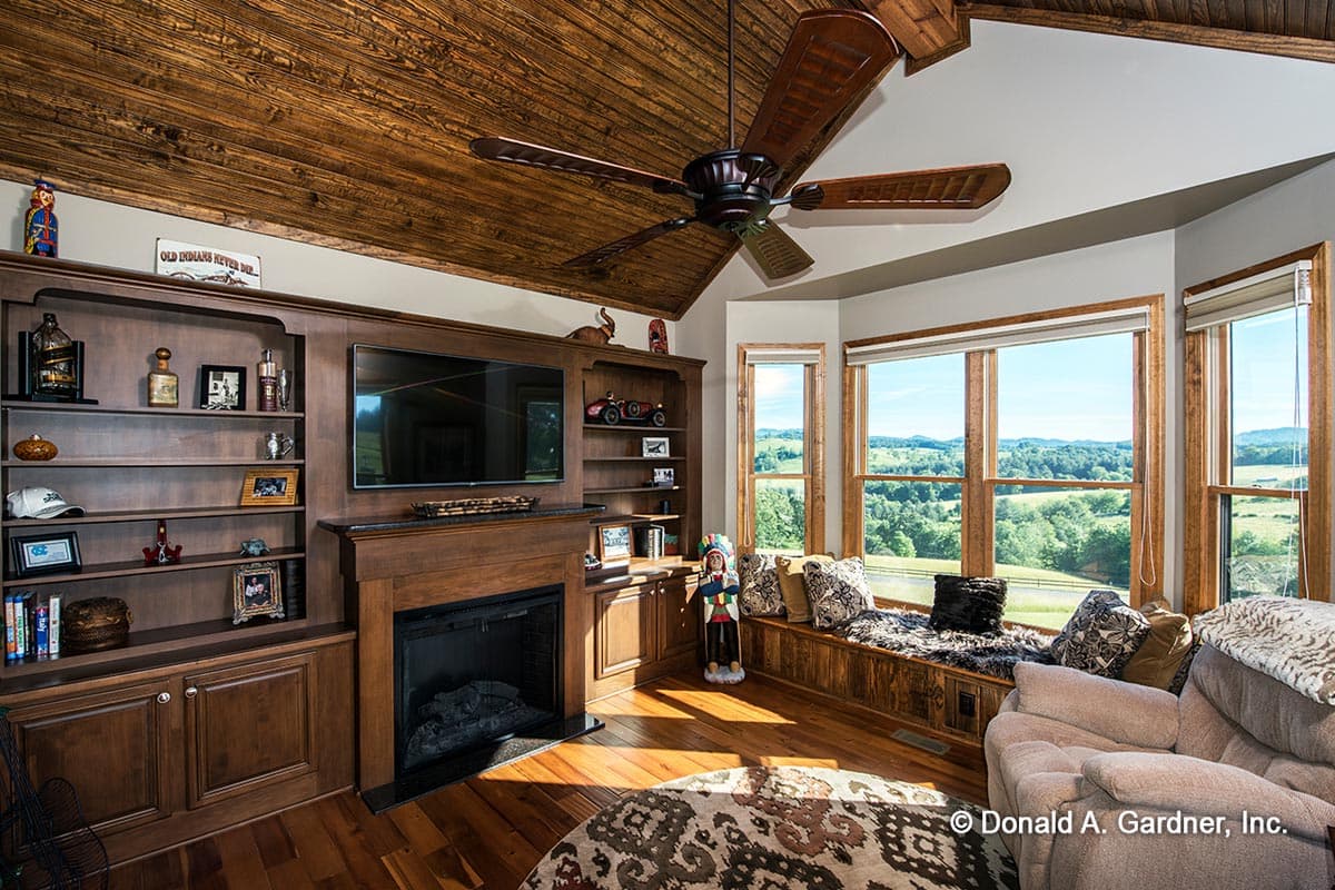 Interior living space with vaulted wood-paneled ceiling, fireplace, built-in shelving, and large windows overlooking a landscape.