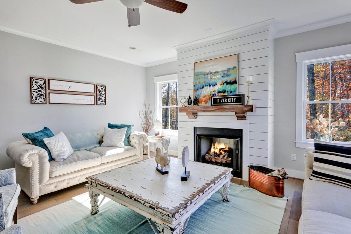Living room interior with fireplace, shiplap accent wall, tufted sofa, large coffee table, and windows overlooking trees.